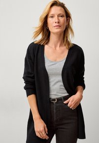 Zwarte cardigan, met open voorkant, lange mouwen, gebreide textuur. Draagt over een grijze top, gecombineerd met donkere spijkerbroeken en een riem. Minimalistisch ontwerp.