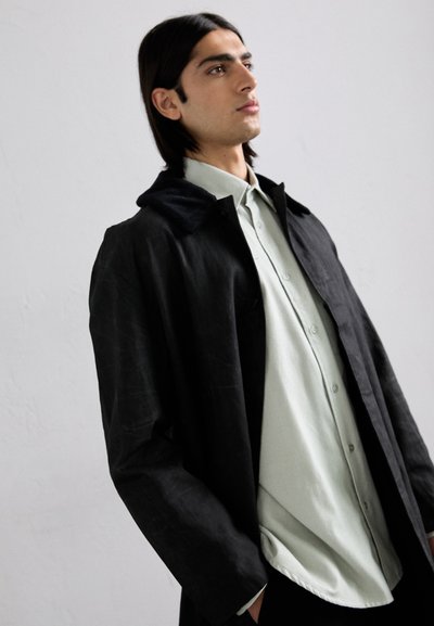 Giacca nera in tessuto morbido e texturizzato, con colletto in velluto a coste, indossata sopra una camicia verde chiaro con bottoni. Design semplice e moderno con una vestibilità rilassata.