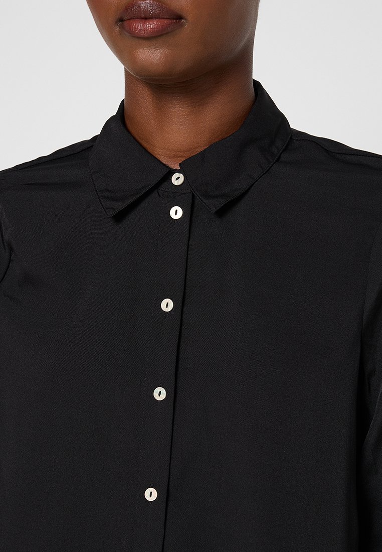 Zwarte button-up shirt met een spitse kraag, voorzien van witte knopen en een gestructureerde stof. Close-up weergave van de kraag en de knoppenlijn.