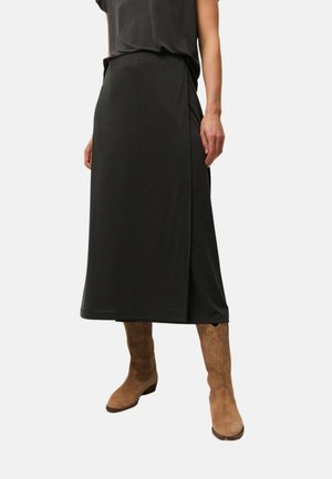 Mujer con falda midi negra con abertura lateral y botas marrones de ante estilo western sobre un fondo liso.