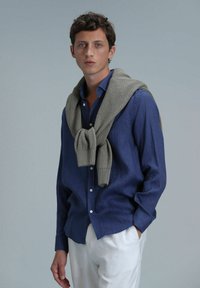 Marineblauw linnen shirt met knopen, gecombineerd met een lichtgrijze gebreide trui over de schouders, gestyled met witte broek.