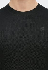 Juodas megztinis su apvaliu kaklu, tekstūruotu avilio raštu ir subtiliu logotipu ant krūtinės, pagamintas iš minkštos medžiagos.