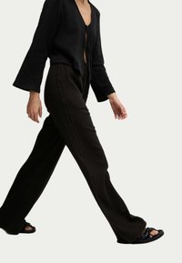 Czarny, fakturowany, krótki top z wiązaniem i dzwonowymi rękawami, w połączeniu z luźnymi, ciemnymi spodniami i płaskimi, czarnymi sandałami.