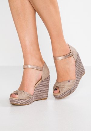 Beige wedge sandals with a knotted fabric upper, ankle strap, and woven jute heel. Open-toe design with a smooth texture and subtle logo detail.