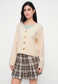 Πλεκτή κοντή ζακέτα σε ροδακινί απόχρωση με V λαιμόκοψη, τέσσερα χρυσά κουμπιά σε σχήμα καρδιάς, φορεμένη πάνω από ένα ανοιχτοπράσινο τοπ και συνδυασμένη με καρό καφέ φούστα.