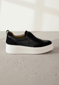 Black slip-on sneakers feature a mix of leather and suede, a thick white platform sole, and decorative stitching across the upper.