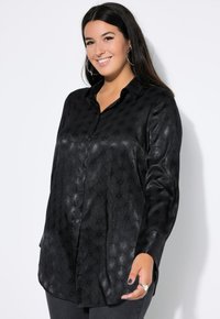 Zwarte button-up shirt met een textuur, reflecterende afwerking en bloemenpatroon. Heeft lange mouwen en een ontspannen pasvorm, gecombineerd met donkere jeans.