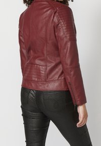 Czerwona skórzana kurtka z prążkowanymi akcentami na ramionach, dopasowany krój i zapięcia na zamek. W zestawie z czarnymi, teksturowanymi spodniami z materiału przypominającego skórę.