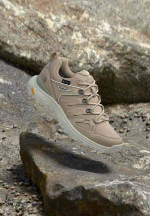 Beige vandresko med snørebånd og Vibram-sål svævende over klippefyldt terræn.