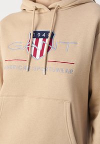 Beige hoodie with a front pocket, featuring an embroidered shield logo in red, white, and navy. Drawstrings with metal tips accentuate the design.