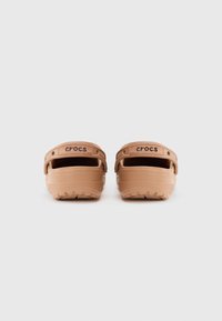 Ett par bruna Crocs-sandaler visade från baksidan med justerbara hälsnören och texturerade sulor på en enkel vit bakgrund.