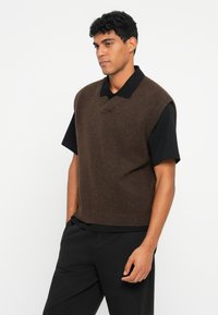 Άνδρας που φοράει ένα σκούρο καφέ αμάνικο πλεκτό γιλέκο πάνω από ένα μαύρο πόλο με κοντά μανίκια και μαύρο παντελόνι, στέκεται με τα χέρια στις τσέπες.