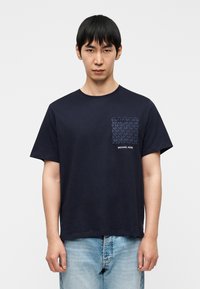 Μπλε βαμβακερό t-shirt με σκούρο χρώμα, με τσέπη στο στήθος με σχέδιο και ετικέτα με κείμενο. Κοντά μανίκια, στρογγ υλή λαιμόκοψη και άνετη γραμμή.