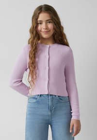 Lila geribde cardigan met lange mouwen, ronde halslijn en zes bijpassende knopen aan de voorkant. Gekleed met lichtblauwe spijkerbroek.
