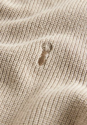 Tessuto lavorato a maglia beige con texture a coste, che presenta un piccolo strappo o foro visibile nel materiale.