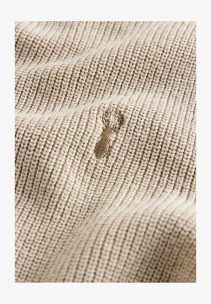 Béžová pletená tkanina s rebrovanou textúrou, ktorú zdobí malá, viditeľná ťahavá značka alebo dierka v materiáli.