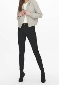 Světle šedá bunda s kapucí a zipem vpředu, pod ní bílá košile, černé úzké džíny a černé boty s podpatkem vystavené na modelce.