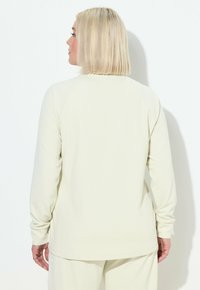Helle beige Langarmbluse aus strukturiertem Rippstoff. Verfügt über Raglanärmel und einen abgerundeten Saum. Rückansicht angezeigt.