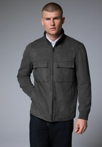 Veste grise dotée d'une fermeture éclair à l'avant, de deux poches poitrine et de poignets côtelés. Fabriquée en tissu doux semblable au daim avec une coupe structurée.