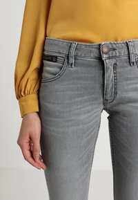 Šedé džíny s úzkým střihem, se předními kapsami, jemným prošíváním a kovovými knoflíky, v kombinaci s žlutou dlouh sleeve blůzou.