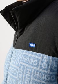 Czarna i jasnoniebieska kurtka zapinana na zamek błyskawiczny z wysokim kołnierzem, z teksturowaną górną częścią i wzorzystą dolną częścią; logo na piersi.