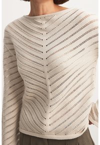 Vaaleabeige neulepaita, jossa on vinottain avoin kudontakuosi, venepääntie ja pitkät hihat; pehmeä tekstuuri, rento istuvuus.