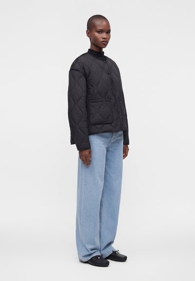 Personne debout, portant une veste noire matelassée, un jean bleu clair à jambes larges et des chaussures noires, sur un fond blanc uni.