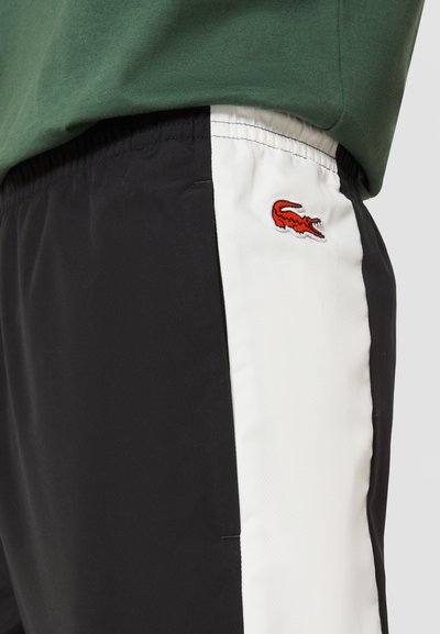 Shorts noirs avec un panneau latéral blanc, arborant un logo de crocodile rouge brodé. Fabriqués en tissu léger, avec une taille élastique pour plus de confort.