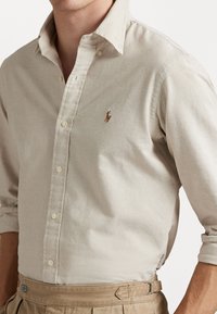 Világos bézs gombos ing klasszikus gallérral, hímzett polo logóval és textúrált anyaggal, khaki redőzött nadrággal párosítva.