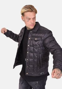 Doudoune noire matelassée avec boutons pression, deux poches poitrine et un éclat texturé. Portée par-dessus une chemise noire avec un logo.