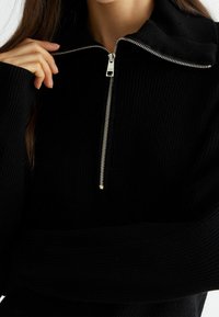 Schwarzer, gerippter Strickpullover mit halbem Reißverschluss, hohem Kragen und silbernem Reißverschlussdetail. Der Stoff hat eine weiche Textur und zeigt volle Ärmel sowie ein figurnahes Design.