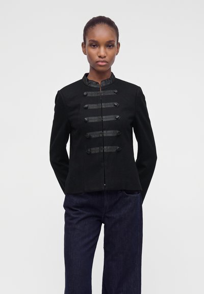 Femme portant une veste noire de style militaire avec des boutons décoratifs et un pantalon bleu foncé, debout devant un fond clair uni.