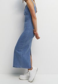 Robe maxi en jean bleu clair, avec un corsage ajusté et une fente latérale. Associée à des baskets blanches pour un look décontracté.