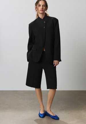Femme debout portant un blazer noir ample et un short noir jusqu'aux genoux avec des ballerines bleu vif sur un fond gris uni.