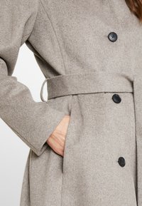 Abrigo de lana gris claro con cintura con cinturón, dos botones y un bolsillo lateral. Se destaca su textura suave y su ajuste estructurado.