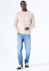 Beige crewneck trui met horizontale strepen, gecombineerd met lichtblauwe jeans en zwarte sneakers met witte veters en crèmekleurige accenten.