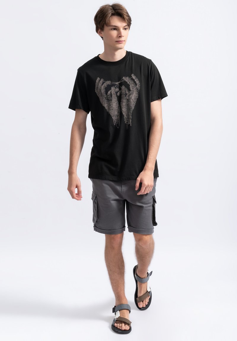 Zwarte katoenen t-shirt met een gedetailleerde handenafbeelding, gecombineerd met grijze cargo shorts en zwarte sandalen met verstelbare banden.