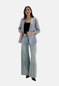 Blazer grigio chiaro con maniche a risvolto, abbinato a jeans azzurri a vita alta e a gamba larga. La modella indossa una maglietta nera e scarpe con tacco nere.