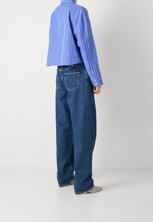 Persoon draagt wijde blauwe spijkerbroek, beige schoenen met een vierkante neus en een cropped blauw gestreept shirt met lange mouwen, gezien van achteren.