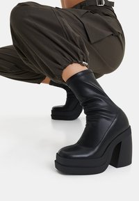Bota de cuero negra hasta el tobillo con un diseño elegante, tacón alto grueso y suela de plataforma. Presenta una textura recogida alrededor del tobillo.