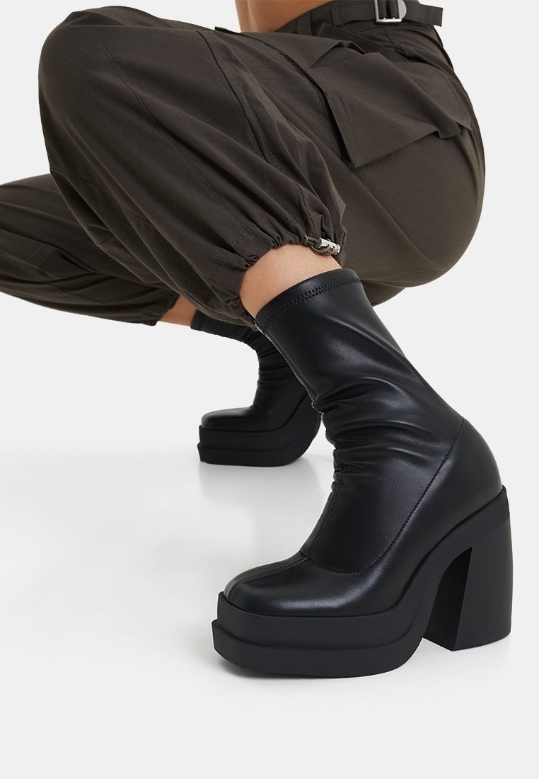 Bota de cuero negra hasta el tobillo con un diseño elegante, tacón alto grueso y suela de plataforma. Presenta una textura recogida alrededor del tobillo.