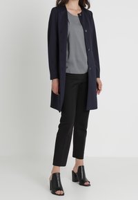 Femme portant un long manteau bleu marine, un chemisier gris à motifs, un pantalon noir cropped, et des mules à talons ouverts noires, se tenant sur un sol blanc.