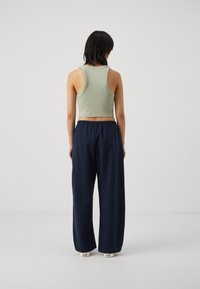 Kvinna med kort mörkt hår som bär en ljusgrön kort ärmad topp, marinblå vida byxor och vita sneakers, stående med ryggen mot oss.