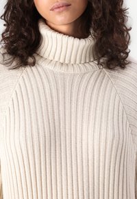 Beige ribbed turtleneck sweater with a textured fabric. Features a high collar and raglan sleeves, highlighting its knitted pattern.