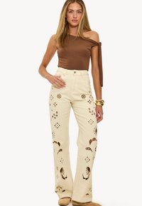 Vaqueros acampanados de color crema con patrones de flores recortados. Presentan una cintura alta y un diseño de cinco bolsillos, hechos de material de mezclilla.