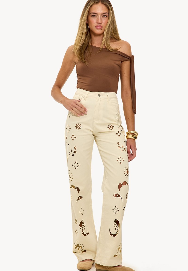 Vaqueros acampanados de color crema con patrones de flores recortados. Presentan una cintura alta y un diseño de cinco bolsillos, hechos de material de mezclilla.