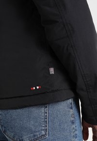Manche et ourlet de veste noire avec petit logo brodé rouge, blanc et noir au-dessus de la poche arrière d'un jean bleu.