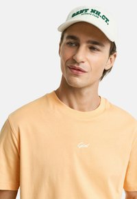 Pfirsichfarbiges Baumwoll-T-Shirt mit Rundhalsausschnitt und kurzen Ärmeln, das ein kleines weißes "Gant"-Logo auf der Brust aufweist. Trägt eine hellfarbige Kappe.