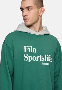Felpa verde con cappuccio grigio, con testo bianco audace "Fila Sportslife. Dal 1911" sul davanti. Tessuto morbido, vestibilità comoda.