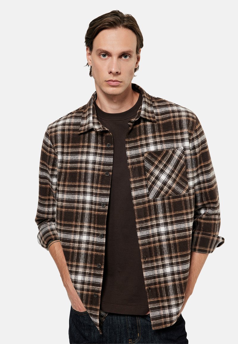 Young man with hands in pockets wearing a brown and white plaid button-up shirt over a dark brown t-shirt.